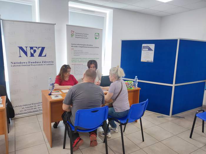 Pokaż zdjęcie: W sali NFZ w Lublinie przy stoliku siedzą cztery osoby. Na stole leżą ulotki. Za stolikiem stoi roll-up z logo Centrum Informacyjno-Doradczego dla Osób z Niepełnosprawnościami.