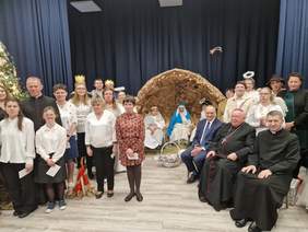 zdjęcie grupowe uczestników wydarzenia na środku z tyłu szopka z postaciami, po bokach uczestnicy w tym z prawej strony osoby duchowne